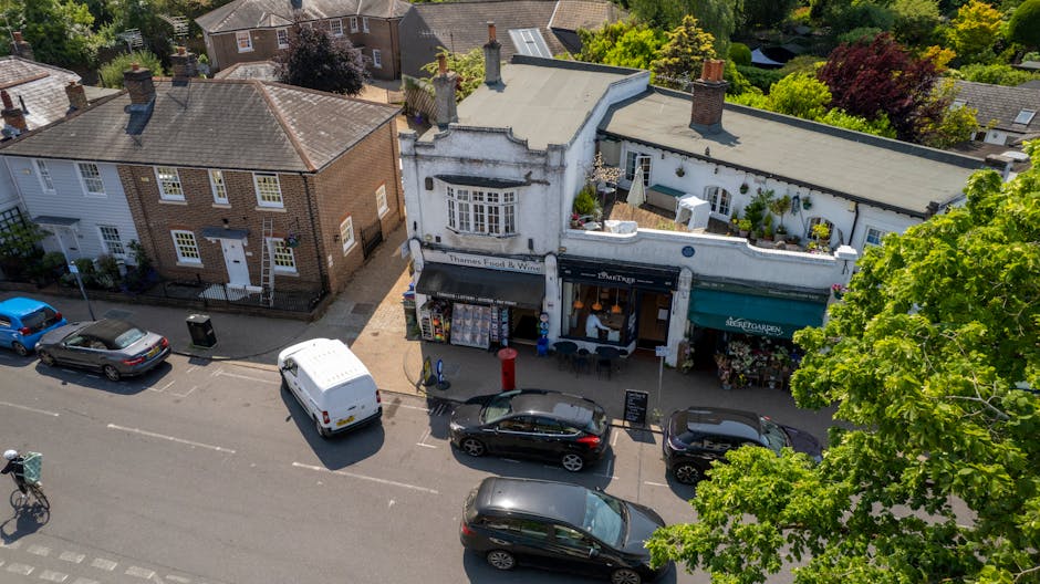 An aerial view of a charming street in Marlow with a white, vintage-style floral shop featuring large shop windows and a black awning, situated alongside modern brick buildings. Parked in front are se