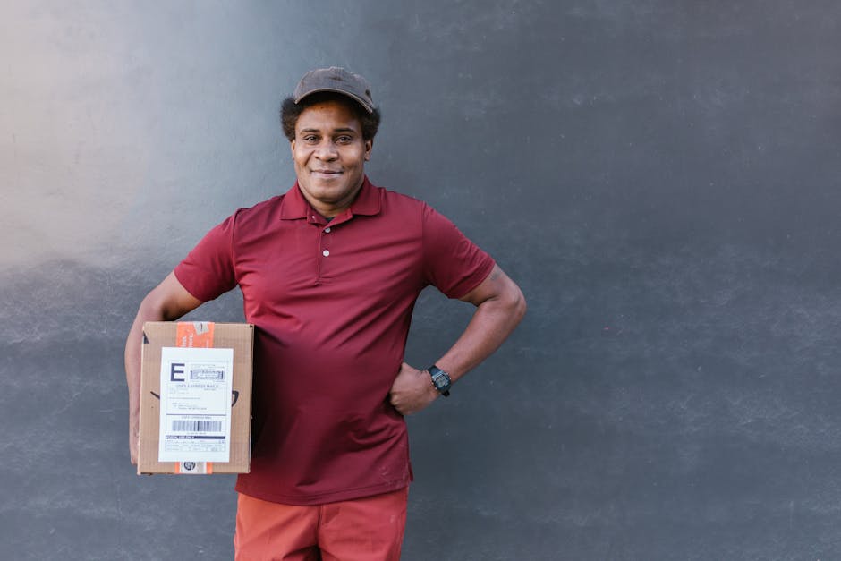 A smiling delivery person with dark skin, wearing a maroon polo shirt, red trousers, and a dark cap, stands against a plain, dark wall. They are holding a small cardboard parcel with a shipping label 