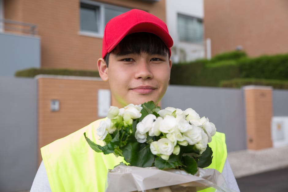 A young male flower delivery courier wearing a bright yellow high-visibility vest and a red cap stands outdoors in front of modern residential buildings. He holds a bouquet of fresh white flowers, inc