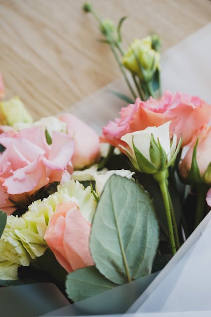 Close-up of a floral arrangement featuring soft pink and cream Lisianthus flowers with delicate, ruffled petals, paired with large, matte green leaves. The bouquet is wrapped in translucent white tiss