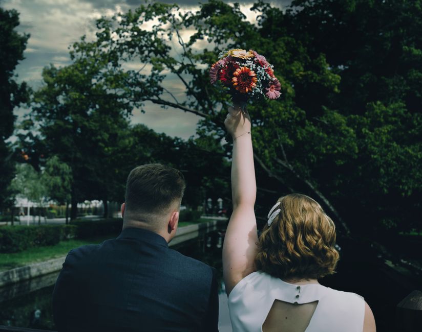 A woman with short, wavy, reddish-brown hair wearing a white sleeveless dress holds a vibrant bouquet of fresh flowers high above her head. The bouquet features a mix of red, orange, pink, and white b