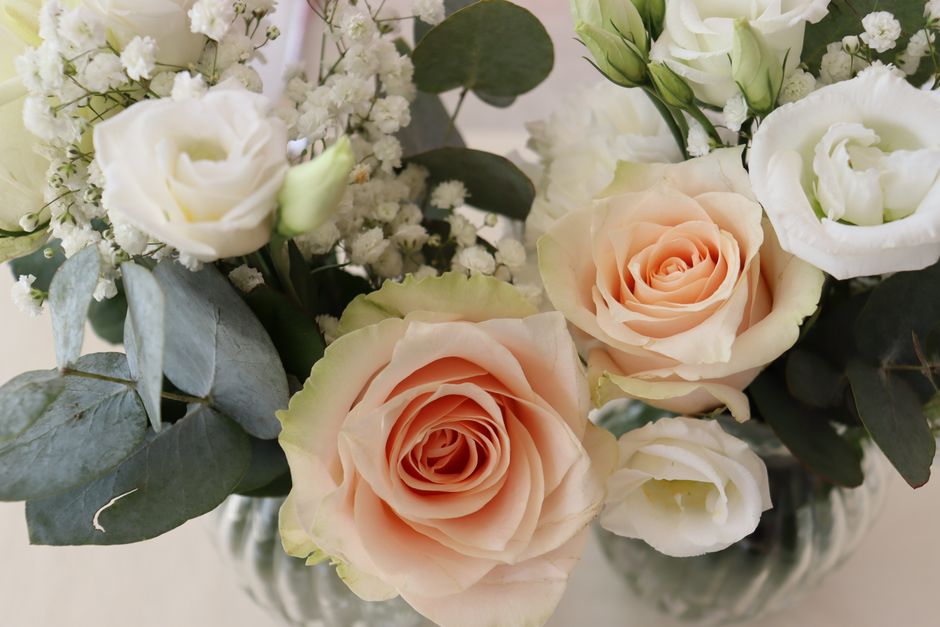 A floral arrangement featuring large, soft peach-colored roses with tightly layered petals, complemented by white lisianthus and delicate white baby's breath, all accented with broad, silvery-green eu